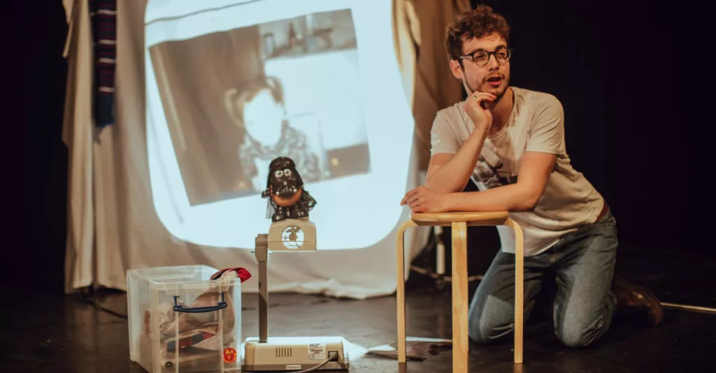 Jack Hunter, One of Two A person leans on a coffee table on stage. There is a projection of an old photograph as the backdrop.