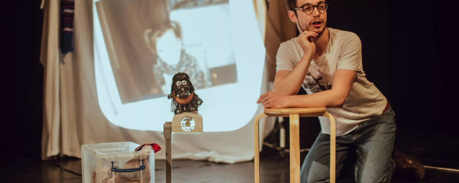 Jack Hunter, One of Two A person leans on a coffee table on stage. There is a projection of an old photograph as the backdrop.